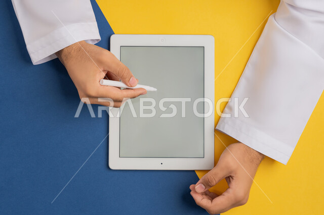 متابعة الدروس التعليمية، عرض شاشة رمادية فارغة، بورتريه علوي عامودي ليدين رجل عربي خليجي إماراتي يقوم بإنجاز المهام المطلوبة عن بعد، شاب سعودي يستخدم جهاز الآيباد، رسم الصور والتصاميم الالكترونية، استعمال قلم التابلت لتدوين ملاحظات خاصة بالعمل، خلفية ملونة