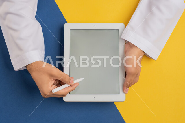 رسم الصور والتصاميم الالكترونية، استخدام قلم التابلت لتدوين ملاحظات خاصة بالعمل، متابعة الدروس التعليمية، عرض شاشة رمادية فارغة، بورتريه علوي عامودي ليدين رجل عربي خليجي إماراتي يقوم بإنجاز المهام المطلوبة عن بعد، شاب سعودي يستعمل جهاز الآيباد، خلفية ملونة