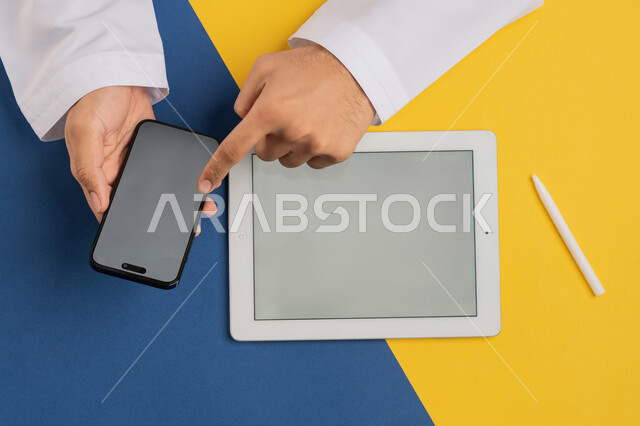 Using modern technical devices in administrative work, distance learning via mobile, benefiting from online shopping applications, vertical top portrait of an Arab Gulf Emirati man holding a mobile phone and a tablet next to him, a Saudi young man transferring files between the two devices, colorful background
