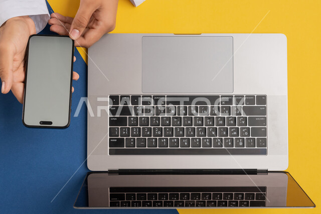 Using modern technical devices in administrative work, marketing and promotion of websites, vertical overhead portrait of an Emirati Gulf Arab man holding a mobile phone, a Saudi young man using a laptop, transferring data and files between the two devices, colorful background