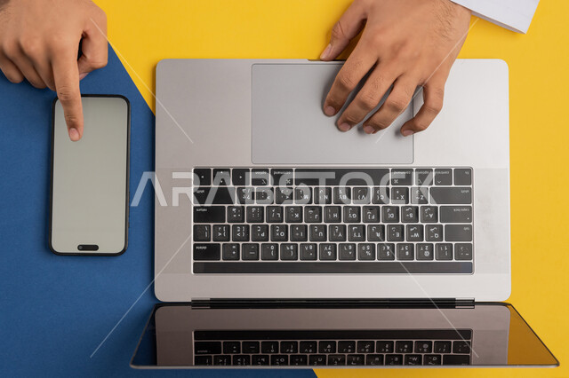 Transferring data and files between two devices, displaying a blank gray screen, using modern technical devices in administrative work, vertical overhead portrait of a man's hands tapping on a mobile phone screen, a Saudi young man using a laptop, marketing and promotion of websites, colorful background