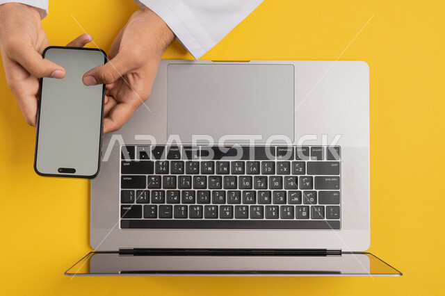 استخدام الاجهزة الحديثة، بورتريه علوي عامودي لرجل عربي خليجي اماراتي يحمل بيده الجوال وبجانبه جهاز اللابتوب، شاب سعودي يقوم بالنقر على شاشة الهاتف المحمول، نقل البيانات والملفات بين الجهازين، عرض شاشة رمادية فارغة، التسويق والترويج للمواقع الالكترونية، خلفية صفراء