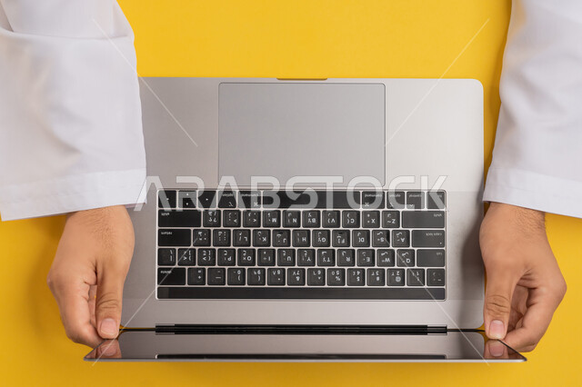 Concept of business management and achieving goals, integrating modern technology into practical life, accomplishing required tasks remotely, vertical top portrait of an Arab Gulf Emirati man working on a laptop, a Saudi young man holding a laptop, enjoying watching through technology, yellow background