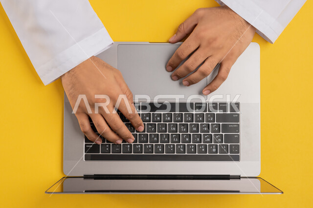 Integrating technology and modern technology into practical life, accomplishing required tasks remotely, the concept of business management and achieving goals, vertical upper portrait of the hands of an Arab Gulf Emirati man using a laptop, a Saudi young man clicking on the laptop keyboard, yellow background