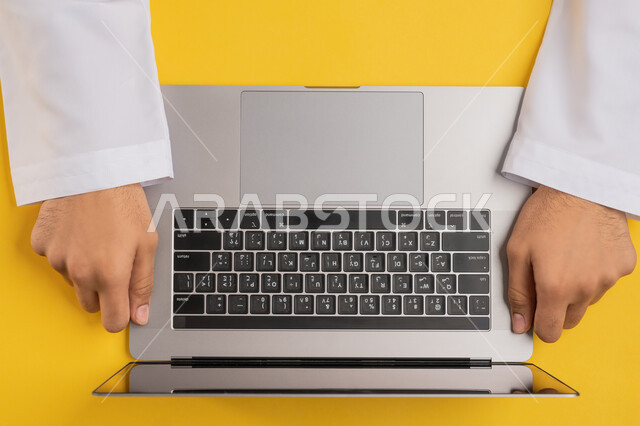 Concept of business management and achieving goals, integrating modern technology into practical life, accomplishing required tasks remotely, vertical top portrait of an Arab Gulf Emirati man working on a laptop, a Saudi young man holding a laptop, enjoying watching through technology, yellow background