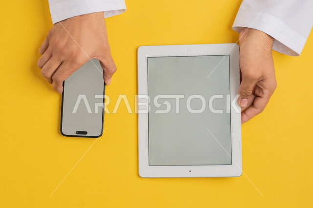 نقل الملفات بين الجهازين، بورتريه علوي عامودي لايدي رجل عربي خليجي اماراتي يحمل الهاتف المحمول وبجانبه جهاز التابلت، شاب سعودي يستخدم التقنية والاجهزة الحديثة، تصفح مواقع التواصل الاجتماعي عبر الانترنت، عرض شاشة رمادية فارغة، خلفية صفراء