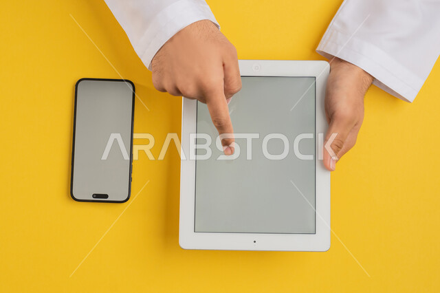 نقل الملفات بين الجهازين، دمج الاجهزة الحديثة في الحياة اليومية والعملية، بورتريه علوي عامودي لايدي رجل عربي خليجي اماراتي يقوم بالنقر على شاشة جهاز التابلت، شاب سعودي يستخدم جهاز الهاتف المحمول، تصفح مواقع التواصل الاجتماعي، عرض شاشة رمادية فارغة، خلفية صفراء