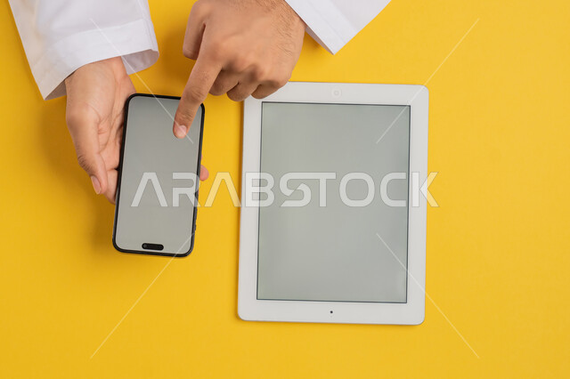 Integrating technology and modern devices into daily and practical life, vertical overhead portrait of an Emirati Gulf Arab man tapping on a mobile phone screen, a Saudi young man using a tablet, browsing social media, displaying a blank gray screen, yellow background