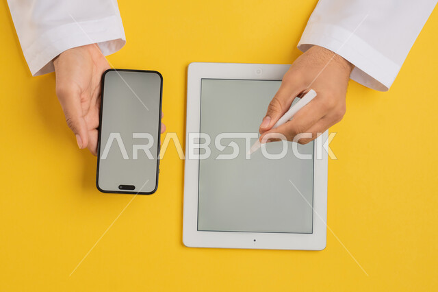 استخدام الاجهزة الحديثة في الاعمال الادارية، بورتريه علوي عامودي لرجل عربي خليجي اماراتي يحمل بيده الجوال وبجانبه جهاز التابلت، شاب سعودي يقوم باستخدام القلم الخاص بالآيباد لتدوين الملاحظات، نقل البيناتات والملفات بين الجهازين، عرض شاشة رمادية فارغة، خلفية صفراء