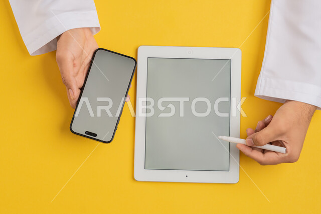 Using modern devices in administrative work, vertical overhead portrait of an Arab Gulf Emirati man holding a mobile phone and a tablet next to him, a Saudi young man using an iPad pen to take notes, transferring data and files between the two devices, displaying a blank gray screen, yellow background