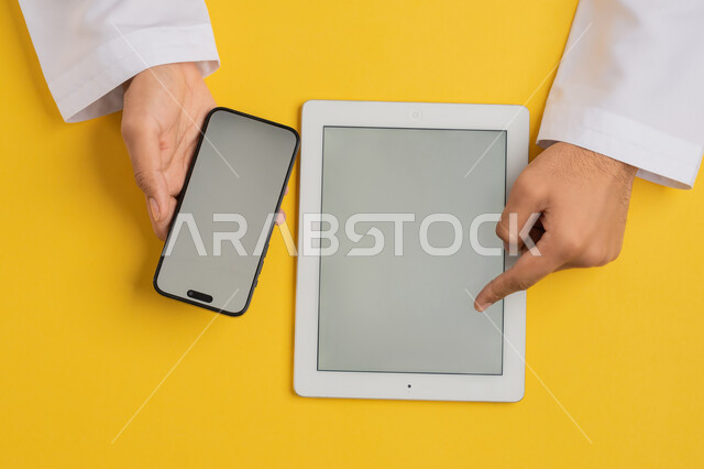 دمج التكنولوجيا الحديثة في الحياة اليومية والعملية، عرض شاشة رمادية فارغة، التسويق الالكتروني للتطبيقات، بورتريه علوي عامودي لايدي رجل عربي خليجي سعودي يقوم بالنقرعلى جهاز التابلت، شاب اماراتي يستخدام الهاتف المحمول، خلفية صفراء