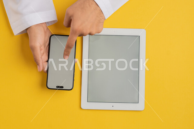 Integrating technology and modern devices into daily and practical life, vertical overhead portrait of an Emirati Gulf Arab man tapping on a mobile phone screen, a Saudi young man using a tablet, browsing social media, displaying a blank gray screen, yellow background