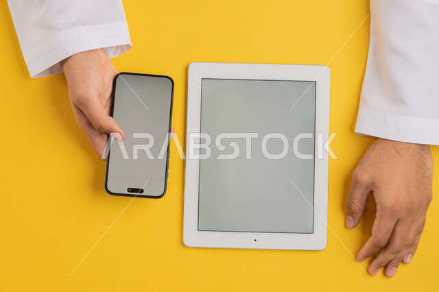 Integrating technology and modern devices into daily and practical life, vertical overhead portrait of an Emirati Gulf Arab man tapping on a mobile phone screen, a Saudi young man using a tablet, browsing social media, displaying a blank gray screen, yellow background