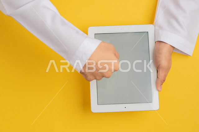 انجاز المهام عن بعد، متابعة الدروس التعليمية، بورتريه علوي عامودي ليد رجل عربي خليجي اماراتي يقوم بالنقر باصبعه السبابة على شاشة جهاز التابلت، شاب سعودي يحمل بيده الآيباد، تصفح مواقع التواصل الاجتماعي، عرض شاشة رمادية فارغة، خلفية صفراء