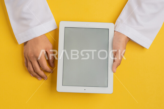استخدام التكنولوجيا والاجهزة الحديثة، بورتريه ليد رجل عربي خليجي اماراتي يحمل بيديه جهاز التابلت، شاب سعودي يستخدم تقنية الايباد، الاستمتاع بمشاهدة الافلام والمسلسلات، عرض شاشة رمادية فارغة، متابعة الدروس التعليمية، خلفية صفراء