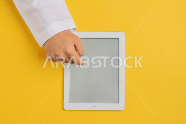 النقر على شاشة رمادية فارغة، دمج التكنولوجيا والتقنيات الحديثة والمتطورة بالعمل، بورتريه علوي عامودي ليد رجل عربي خليجي إماراتي يستخدم التابليت، شاب سعودي يقوم  بتصفح مواقع التواصل الاجتماعي، انجاز المهام المطلوبة عن بعد، خلفية صفراء