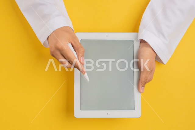 Follow educational lessons, blank gray screen display, vertical overhead portrait of hand of Arab Gulf Emirati man using tablet pen to take notes for work, completing required tasks remotely, Saudi young man holding iPad in hand, drawing pictures and electronic designs, yellow background