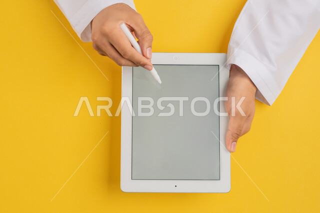 Follow educational lessons, blank gray screen display, vertical overhead portrait of hand of Arab Gulf Emirati man using tablet pen to take notes for work, completing required tasks remotely, Saudi young man holding iPad in hand, drawing pictures and electronic designs, yellow background