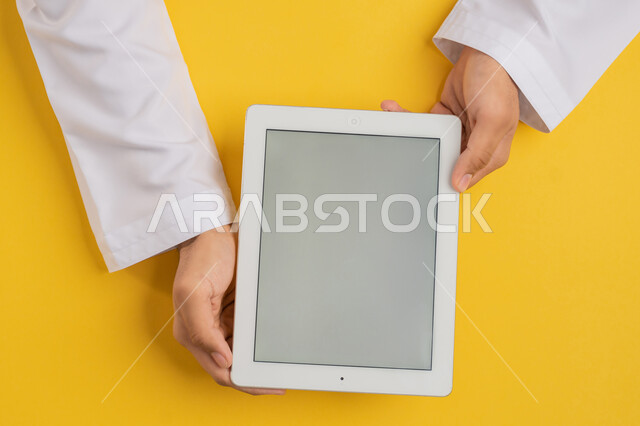 استخدام التكنولوجيا والاجهزة الحديثة، بورتريه ليد رجل عربي خليجي اماراتي يحمل بيديه جهاز التابلت، شاب سعودي يستخدم تقنية الايباد، الاستمتاع بمشاهدة الافلام والمسلسلات، عرض شاشة رمادية فارغة، متابعة الدروس التعليمية، خلفية صفراء