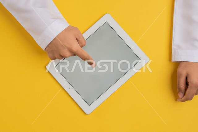 Blank gray screen, completing tasks remotely, following educational lessons, vertical overhead portrait of hand of Arab Gulf Emirati man tapping his index finger on iPad screen, Saudi young man using tablet, browsing social media, locating a specific location, yellow background
