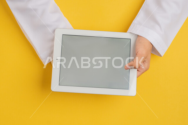 Using modern technology and devices, portrait of a hand of an Arab Gulf Emirati man holding a tablet, a Saudi young man using iPad technology, enjoying watching movies and series, displaying a blank gray screen, following educational lessons, yellow background