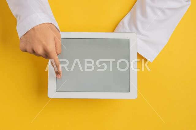 Completing tasks remotely, following educational lessons, vertical overhead portrait of hand of Arab Gulf Emirati man tapping his index finger on tablet screen, Saudi young man holding iPad in hand, browsing social media, blank gray screen display, yellow background