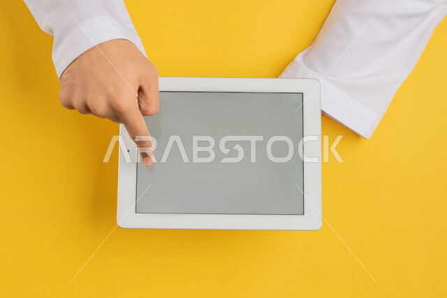 انجاز المهام عن بعد، متابعة الدروس التعليمية، بورتريه علوي عامودي ليد رجل عربي خليجي اماراتي يقوم بالنقر باصبعه السبابة على شاشة جهاز التابلت، شاب سعودي يحمل بيده الآيباد، تصفح مواقع التواصل الاجتماعي، عرض شاشة رمادية فارغة، خلفية صفراء