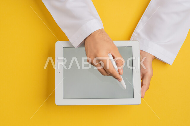 Using tablet pen to take notes for work, following educational lessons, showing blank gray screen, vertical top portrait of hands of Arab Gulf Emirati man completing required tasks remotely, Saudi young man using iPad, drawing pictures and electronic designs, yellow background