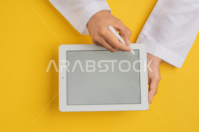 Drawing pictures and electronic designs, using a tablet pen to take notes for work, following educational lessons, displaying a blank gray screen, vertical top portrait of the hands of an Arab Gulf Emirati man completing required tasks remotely, a Saudi young man using an iPad, yellow background