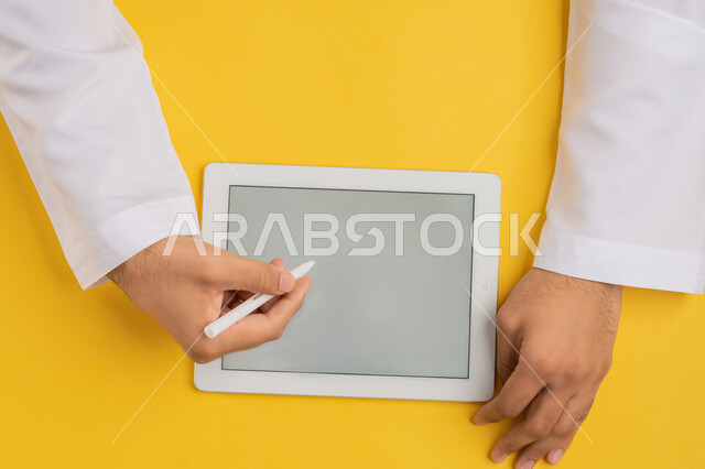 Using tablet pen to take notes for work, following educational lessons, showing blank gray screen, vertical top portrait of hands of Arab Gulf Emirati man completing required tasks remotely, Saudi young man using iPad, drawing pictures and electronic designs, yellow background
