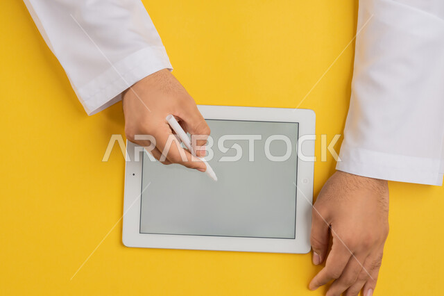 Blank gray screen display, using tablet pen to take notes for work, following educational lessons, vertical overhead portrait of hands of an Arab Gulf Emirati man completing required tasks remotely, Saudi young man using iPad, drawing pictures and electronic designs, yellow background