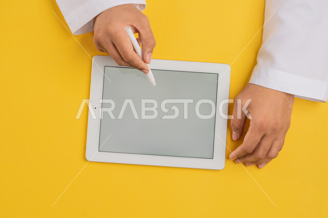 Using tablet pen to take notes for work, following educational lessons, showing blank gray screen, vertical top portrait of hands of Arab Gulf Emirati man completing required tasks remotely, Saudi young man using iPad, drawing pictures and electronic designs, yellow background