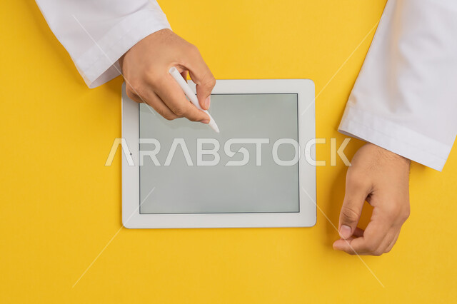 Using tablet pen to take notes for work, following educational lessons, showing blank gray screen, vertical top portrait of hands of Arab Gulf Emirati man completing required tasks remotely, Saudi young man using iPad, drawing pictures and electronic designs, yellow background