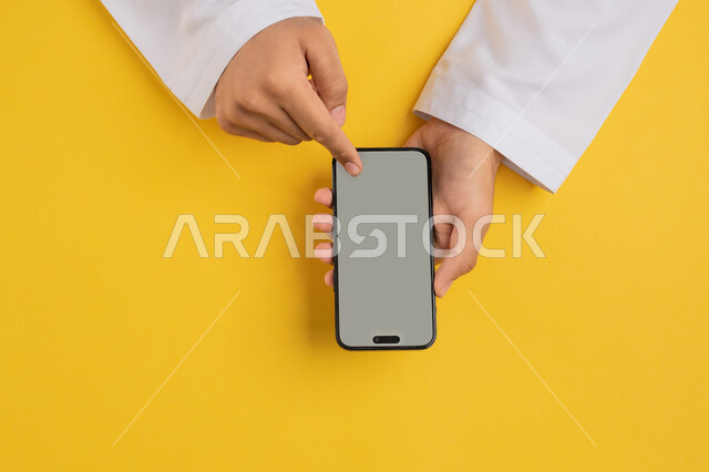 Using technology, technology and modern devices, vertical portrait of an Arab Gulf Emirati man's hand tapping on a mobile phone screen, a Saudi young man holding a mobile phone in his hand, browsing social media sites, specifying a specific location, displaying a blank gray screen, yellow background
