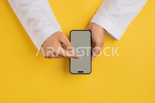 Using technology, technology and modern devices, vertical portrait of an Arab Gulf Emirati man's hand tapping on a mobile phone screen, a Saudi young man holding a mobile phone in his hand, browsing social media sites, specifying a specific location, displaying a blank gray screen, yellow background