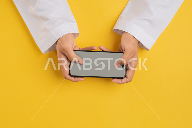 اللعب في الالعاب الالكترونية الترفيهية، دمج التسلية بالتقنية والتكنولوجيا، بورتريه علوي عامودي ليد رجل عربي خليجي اماراتي يحمل بيده الجوال، شاب سعودي يقوم بالنقر باصبعيه الابهام على شاشة الهاتف المحمول، تحديد موقع معين، عرض شاشة رمادية فارغة، خلفية صفراء