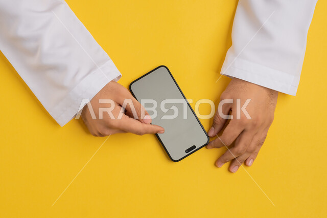 Development of technology, technology and modern devices, vertical portrait of an Arab Gulf Emirati man's hand tapping on a mobile phone screen, a Saudi young man using a mobile phone, browsing social media sites, displaying a blank gray screen, yellow background