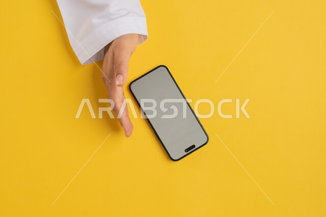 Using technology and modern devices, vertical overhead portrait of an Arab Gulf Emirati man using a mobile phone, a Saudi young man putting his hand behind the mobile phone with gestures to block vision and viewing, showing a blank gray screen, yellow background