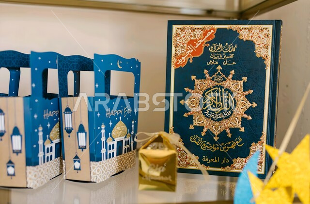 طقوس الاحتفال بالأعياد الإسلامية في المملكة العربية السعودية، تعليق الزينة والأضواء لاستقبال العيد، صورة مقربة للقرآن الكريم بجانبه عدد من علب التوزيعات الملونة، صناديق الحلويات الشهية للأطفال والزوار، إكرام الضيوف وتوزيع الهدايا في المناسبات السعيدة