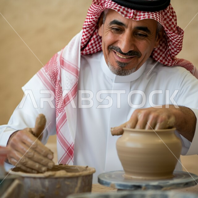 صنع الجرار الفخارية المزخرفة، أواني خزفية مصنوعة من الطين، احتراف العمل في صناعة الخزفيات، حرف وصناعات يدوية شعبية من التراث السعودي العريق، رجل عربي خليجي سعودي يرتدي الثوب التقليدي والشماغ يقوم بصنع إناء من الفخار، أعمال محلية وطنية بأيدي سعودية