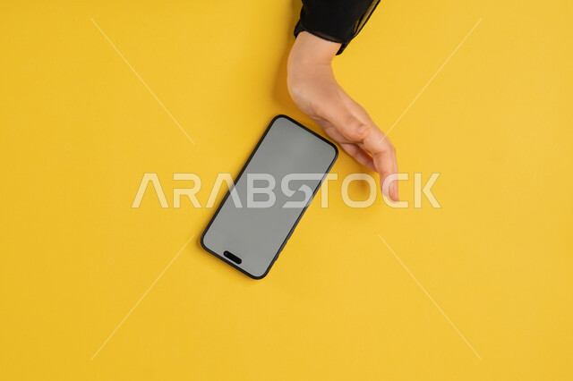 Explaining the steps of working on a mobile phone, using modern technology and techniques, vertical upper portrait of the hands of an Arab Gulf Emirati woman holding a mobile phone, a young Saudi woman wearing an abaya browsing social media sites, displayed on a blank gray screen, yellow background