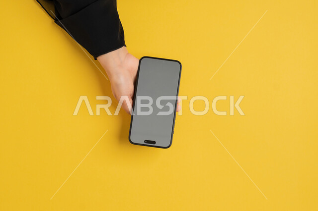 استخدام التكنولوجيا والتقنيات الحديثة، بورتريه علوي عامودي ليدين إمرأة عربية خليجية إماراتية تحمل الهاتف المحمول، شابة سعودية ترتدي العباءة تقوم بتصفح مواقع التواصل الاجتماعي، عرض على شاشة رمادية فارغة، شرح خطوات العمل على الموبايل، خلفية صفراء
