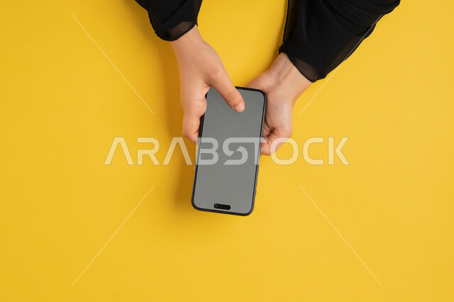 دمج التكنولوجيا والتقنيات الحديثة المتطورة بالعمل، بورتريه علوي عامودي ليدين إمرأة عربية خليجية إماراتية تحمل الهاتف المحمول، شابة سعودية ترتدي العباءة تقوم بتصفح مواقع التواصل الاجتماعي، القيام بعمليات التسويق الالكتروني، عرض شاشة رمادية فارغة، خلفية صفراء