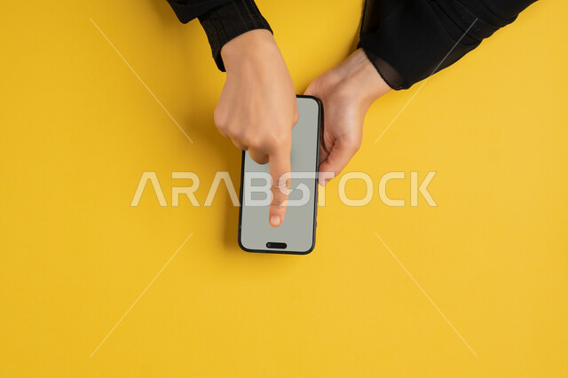 تصفح مواقع التواصل الاجتماعي، النقر على شاشة رمادية فارغة، بورتريه علوي عامودي ليدين إمرأة عربية خليجية إماراتية تقوم بحمل الهاتف المحمول، شابة سعودية ترتدي العباءة تقوم بمتابعة الدروس التعليمية أونلاين، البيع والشراء عبر الانترنت، خلفية صفراء