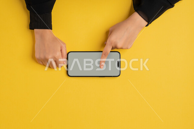 البيع والشراء عبر الانترنت، تصفح مواقع التواصل الاجتماعي، النقر على شاشة رمادية فارغة، بورتريه علوي عامودي ليدين إمرأة عربية خليجية إماراتية تقوم بحمل الهاتف المحمول، شابة سعودية ترتدي العباءة تقوم بمتابعة الدروس التعليمية أونلاين، خلفية صفراء