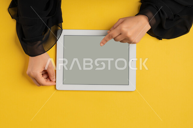 القيام بعمليات التسويق الالكتروني، النقر على شاشة رمادية فارغة، دمج التكنولوجيا والتقنيات الحديثة والمتطورة بالعمل، بورتريه علوي عامودي ليدين إمرأة عربية خليجية إماراتية تحمل التابليت، شابة سعودية ترتدي العباءة تقوم بتصفح مواقع التواصل الاجتماعي، خلفية صفراء
