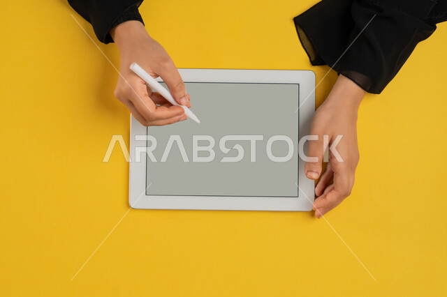 كتابة وتدوين الملاحظات باستخدام قلم التابليت الخاص، بورتريه علوي عامودي ليدين إمرأة عربية خليجية إماراتية تقوم بإنجاز المهام المطلوبة عن بعد، شابة سعودية ترتدي العباءة تستعمل جهاز الآيباد، رسم الصور والتصاميم الالكترونية، عرض شاشة رمادية فارغة، خلفية صفراء