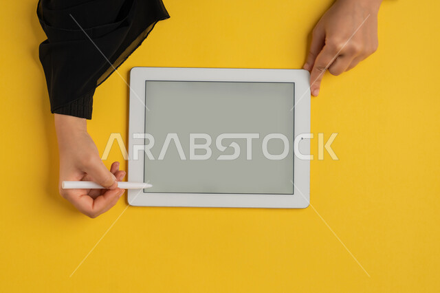 عرض شاشة رمادية فارغة، بورتريه علوي عامودي ليدين إمرأة عربية خليجية إماراتية تقوم بإنجاز المهام المطلوبة عن بعد، شابة سعودية ترتدي العباءة تستعمل جهاز الآيباد، رسم الصور والتصاميم الالكترونية، استخدام قلم التابلت لتدوين ملاحظات خاصة بالعمل، خلفية صفراء