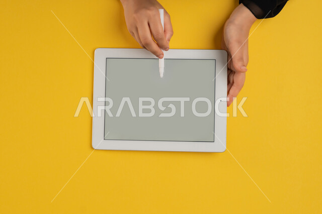 Using tablet pen to take notes for work, blank gray screen view, vertical overhead portrait of hands of an Arab Gulf Emirati woman completing tasks remotely, young Saudi woman wearing abaya using iPad, drawing pictures and electronic designs, colorful background