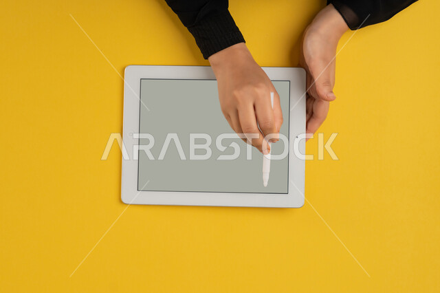 رسم الصور والتصاميم الالكترونية، استخدام قلم التابلت لتدوين ملاحظات خاصة بالعمل، عرض شاشة رمادية فارغة، بورتريه علوي عامودي ليدين إمرأة عربية خليجية إماراتية تقوم بإنجاز المهام المطلوبة عن بعد، شابة سعودية ترتدي العباءة تستعمل جهاز الآيباد، خلفية صفراء